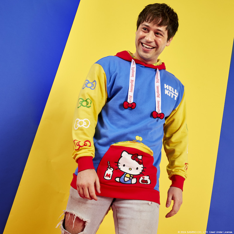 Man wearing the red, blue, and yellow color blocked Hello Kitty 50th Anniversary Unisex Hoodie and standing against a blue and yellow background